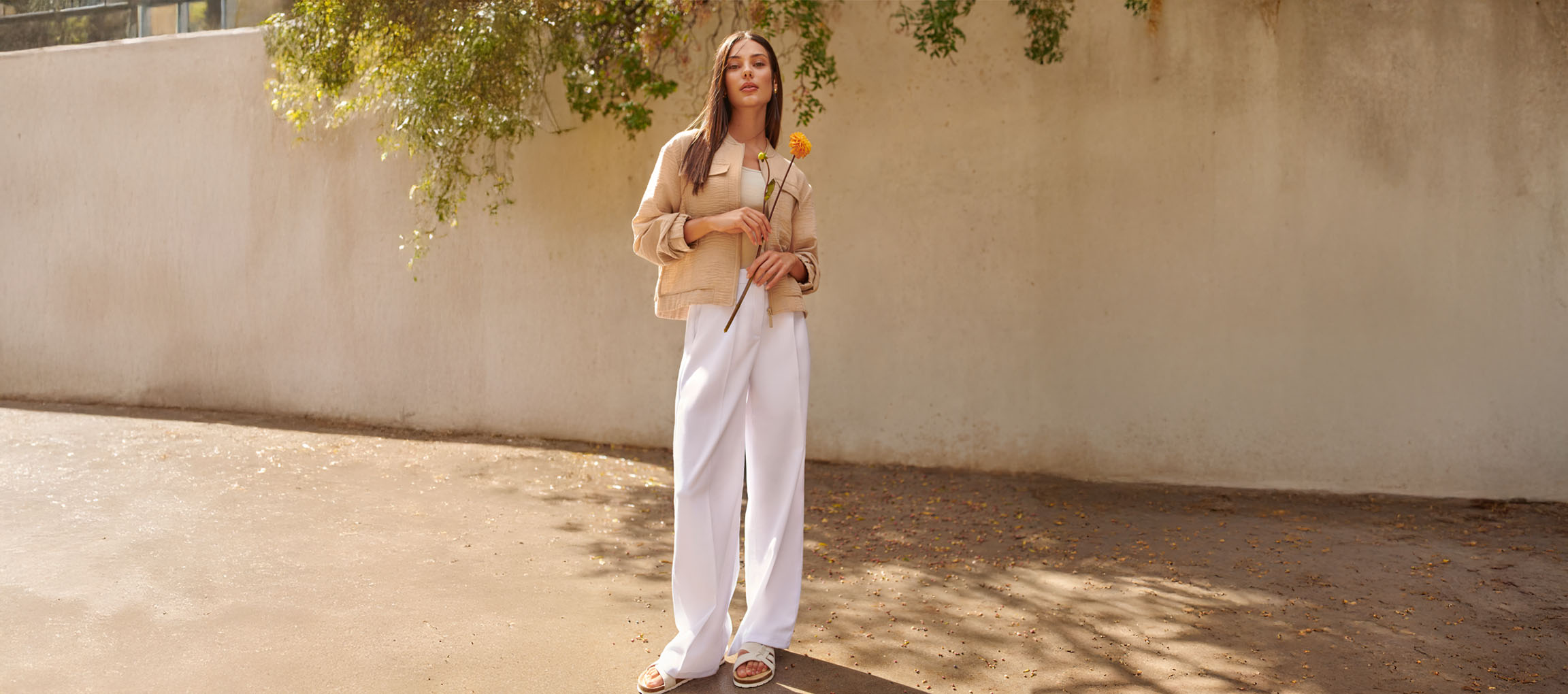 Femme en veste beige, pantalon blanc et sandales, tenant une fleur jaune.