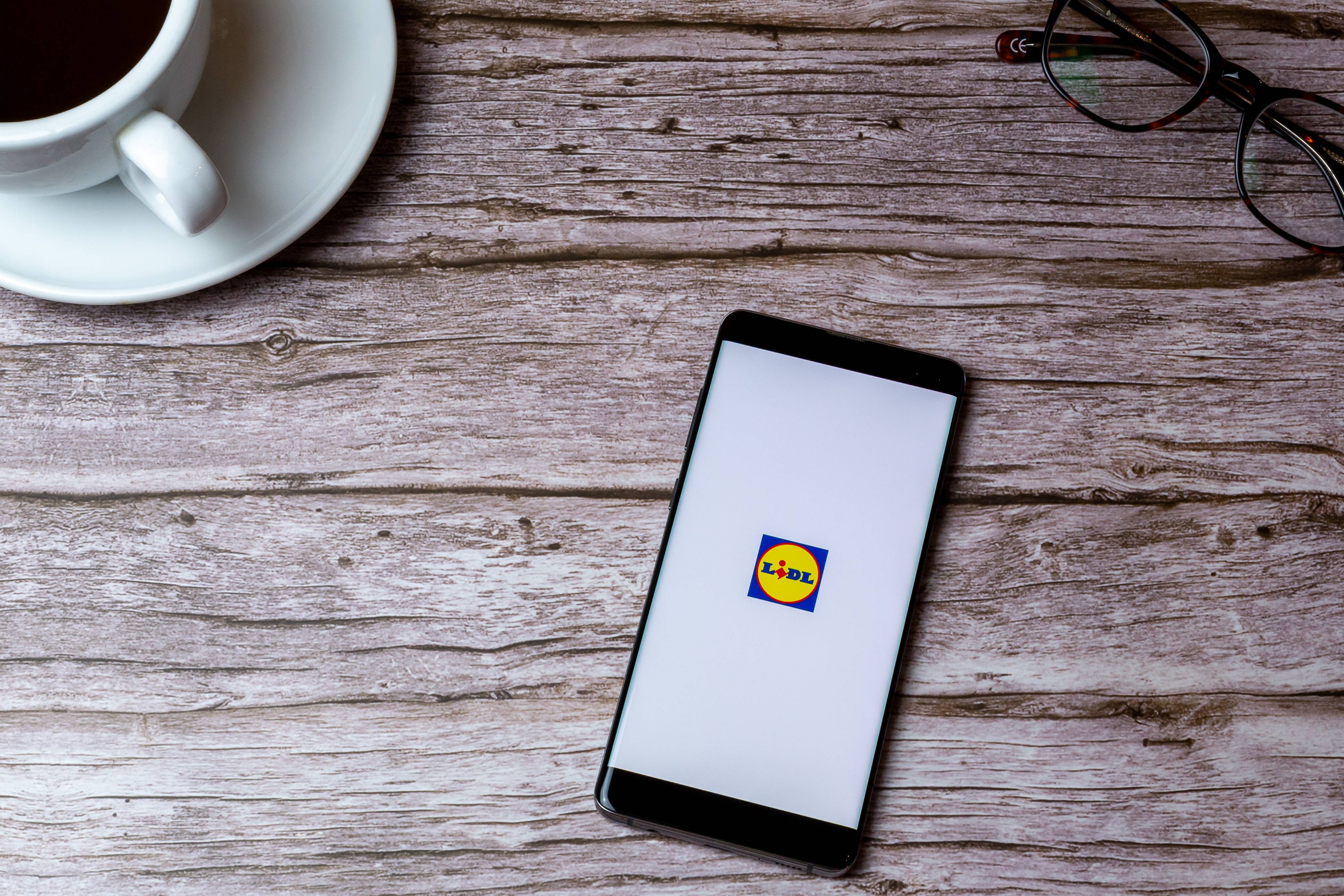 Un smartphone affichant le logo Lidl, une tasse de café et des lunettes sur une table en bois.