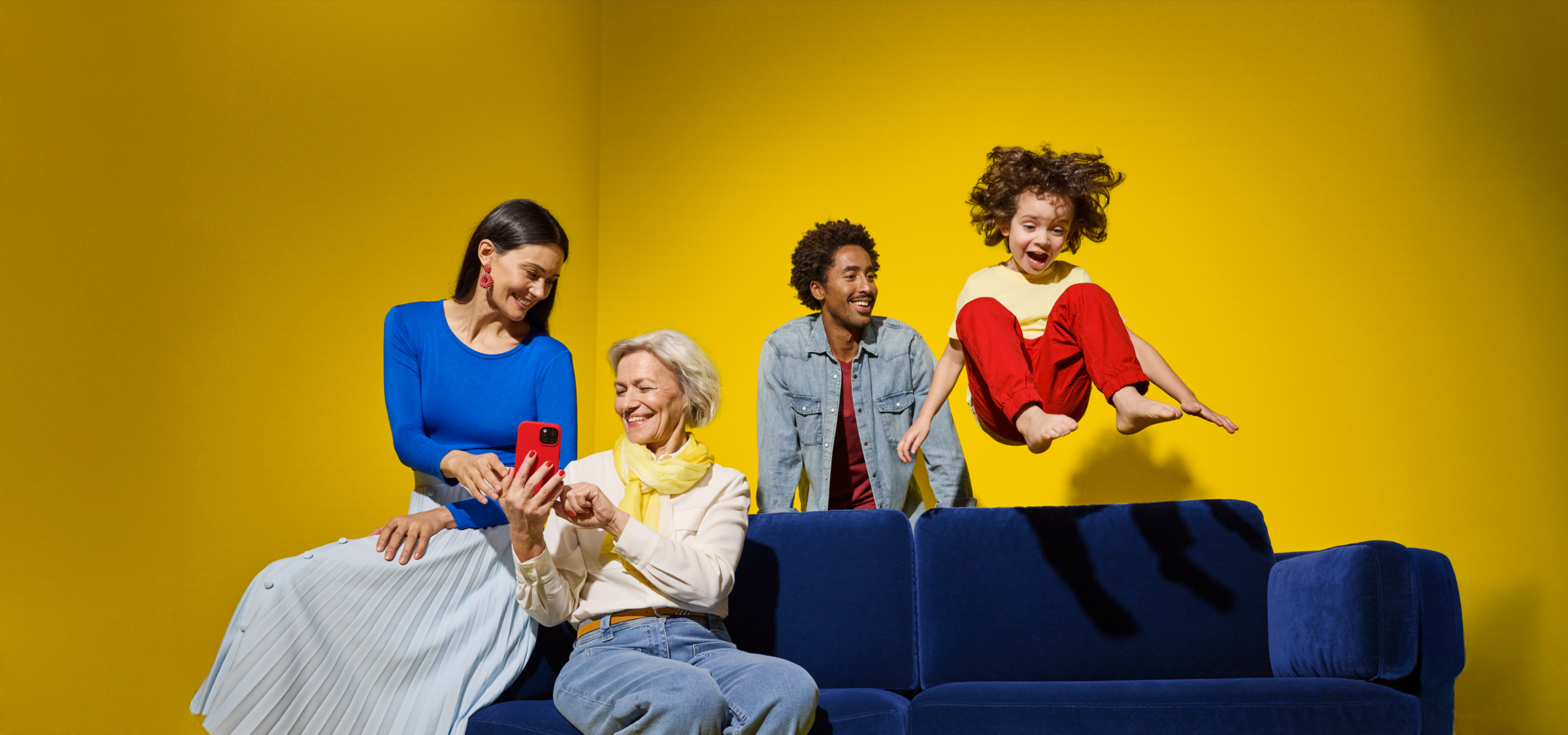 Une famille intergénérationnelle souriante interagit avec un smartphone sur un canapé bleu.