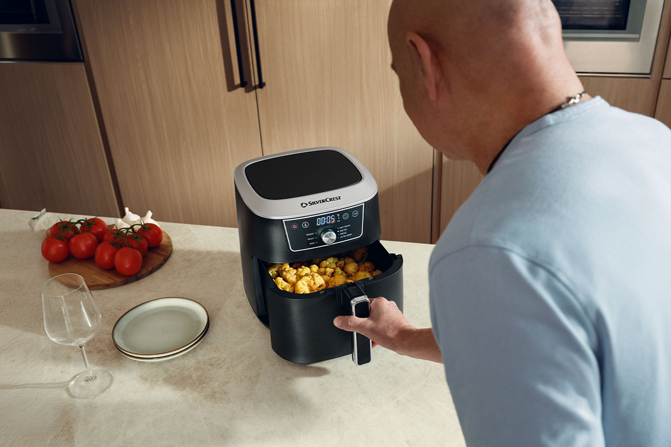 Un homme cuisine avec une friteuse à air dans une cuisine, avec des tomates et des assiettes.