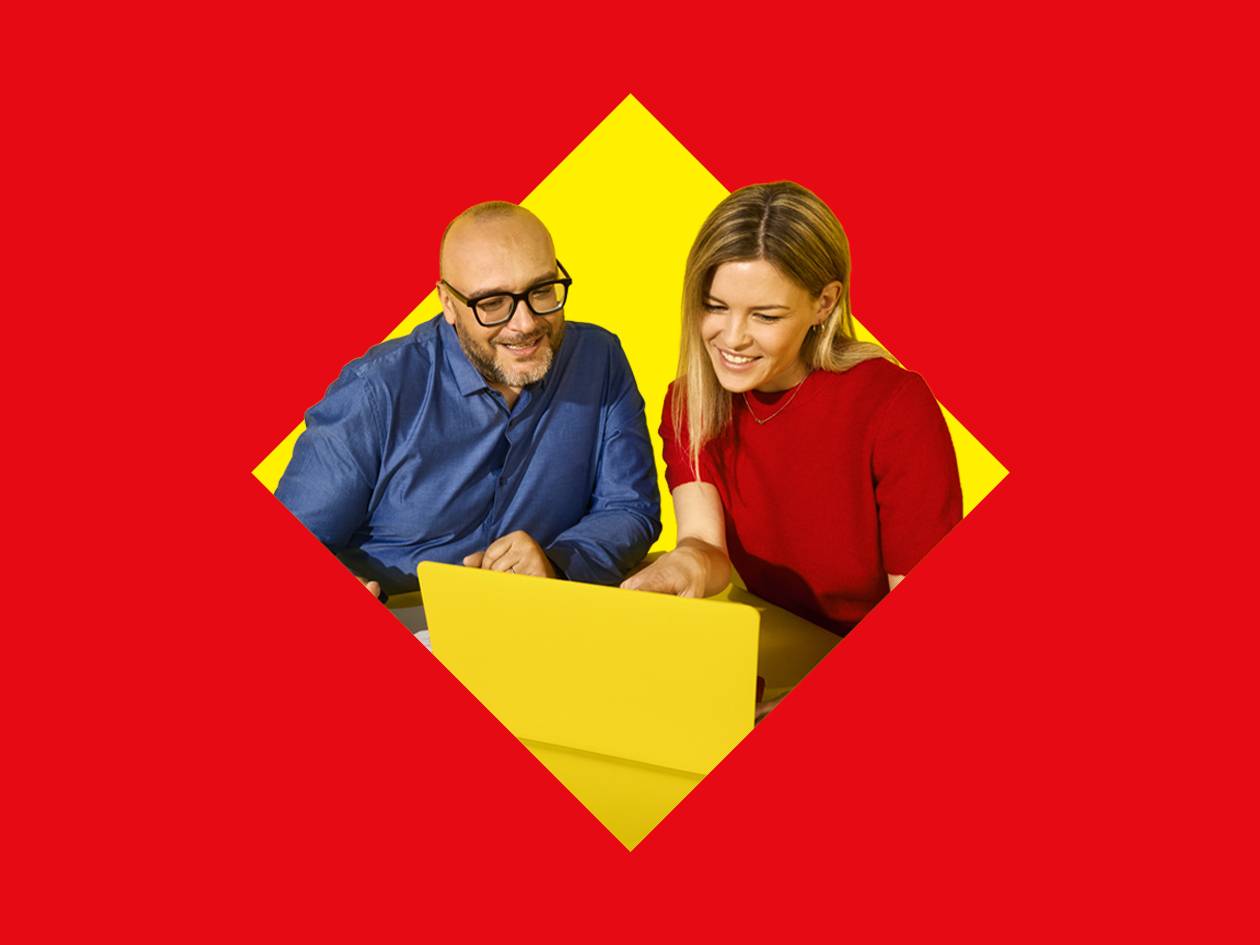Deux personnes souriantes regardant un ordinateur portable jaune sur fond rouge et jaune.