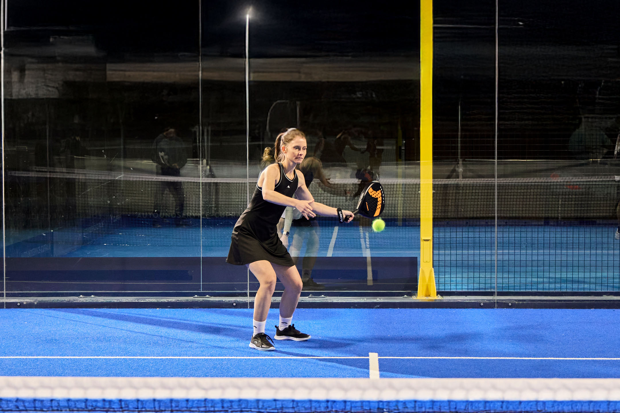 Femme en tenue de sport jouant au padel sur un court bleu avec raquette et balle.